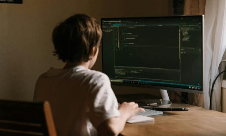 A photo of a person sitting at a computer, working on cybersecurity. The person is wearing a headset and looking focused. The computer screen shows a complex network of data and code